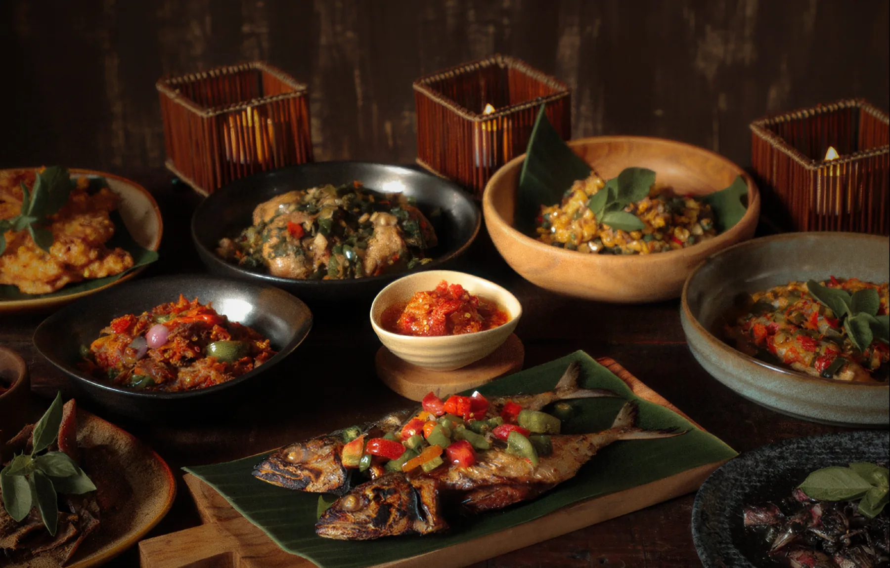 A table of Indonesian dishes in dark bowls: grilled fish with sambal, curries and sides
