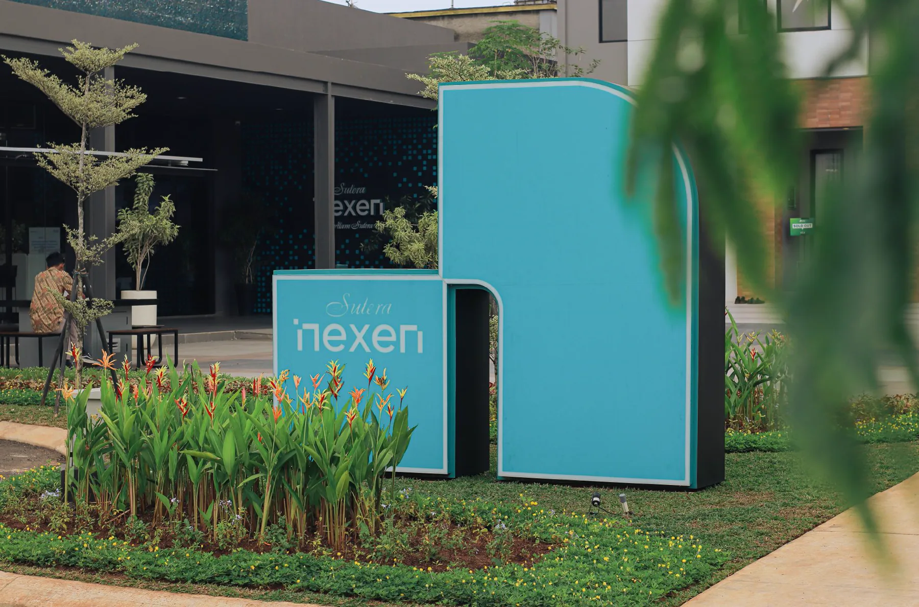 Teal Nexen logo sign and a tall teal N-shaped marker among tropical plants in front of a show house
